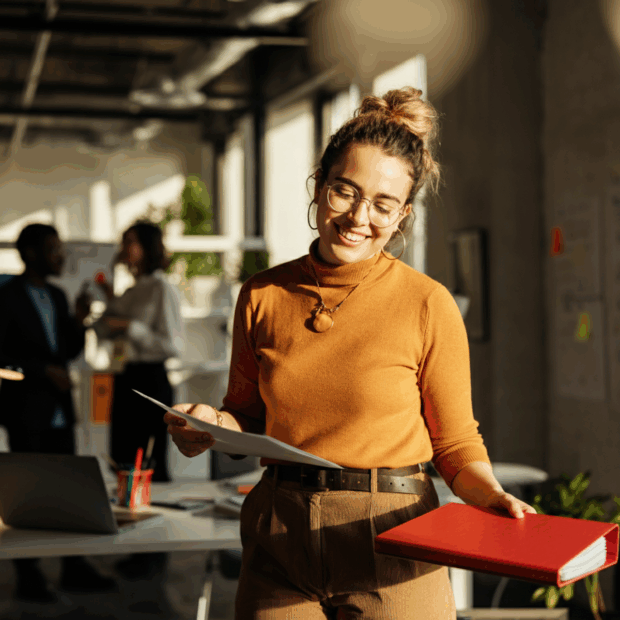 Frau mit Brille und braunem Pullover hält Dokumente und einen roten Ordner in einem modernen Büro.